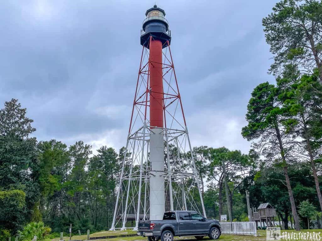 Crooked River Lighthouse in Carrabelle, Florida. The lighthouse is tall and slender, with a red and white color scheme. It consists of a white, tapered lower section supported by a lattice of metal beams, transitioning to a red upper section beneath the lantern room, which is painted black. The lighthouse is surrounded by a dense growth of green trees, suggesting a forested area. In front of the lighthouse, there is a black pickup truck parked on a grassy area.