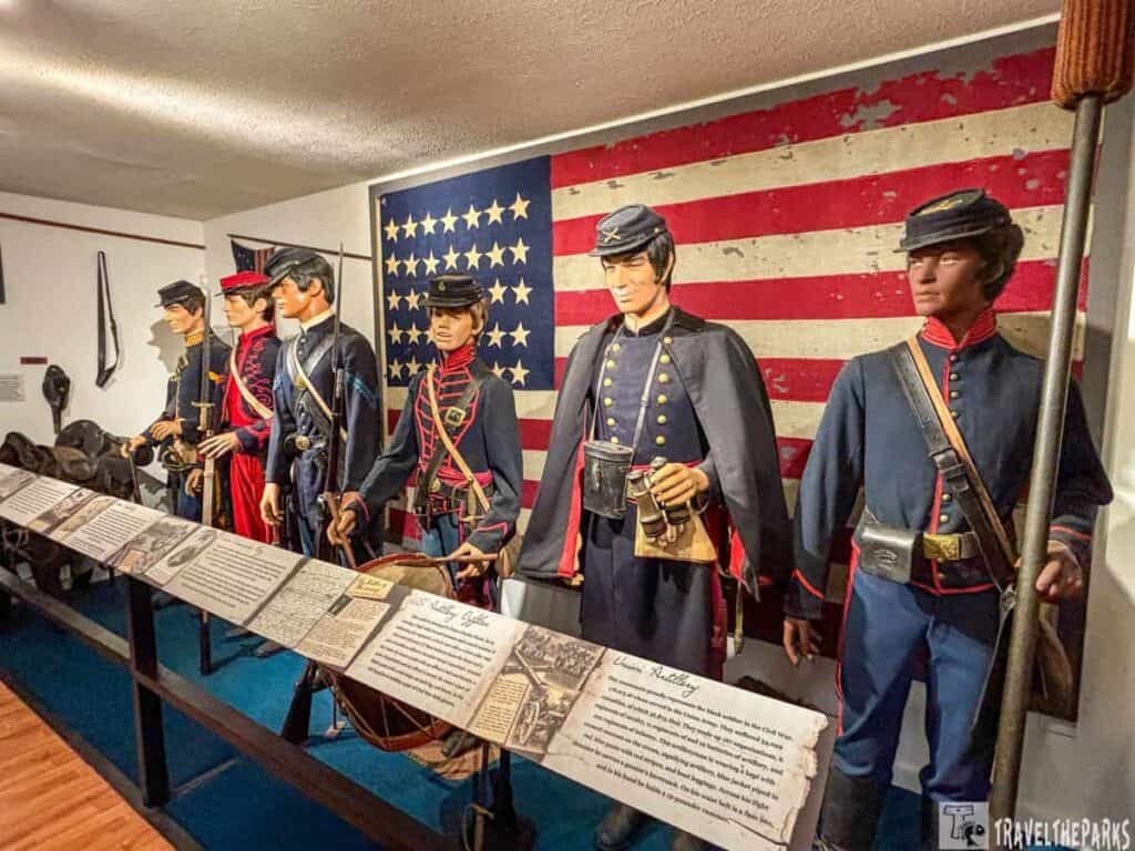 Drummer Boy museum exhibit featuring six mannequin figures dressed in Union military uniforms from the American Civil War era. The mannequins are positioned in a row, each wearing period-appropriate attire, including hats and accessories like drums, swords, and bags. Behind the mannequins, a large American flag is displayed, its stars and stripes visible but slightly worn and faded. The mannequins are set on a blue carpeted platform, and in front of them is a long horizontal display panel featuring informational texts and photographs related to the exhibit. The exhibit is housed within a room with white walls and a wooden floor. The lighting is warm, casting a natural glow over the scene.