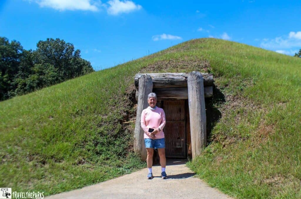 Person standing in front of a grassy earthen mound with a wooden doorway.