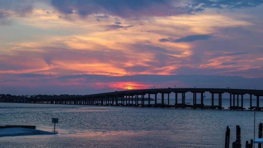 a vibrant sunset over a wide expanse of water, likely depicting the Emerald Coast. A long, elevated bridge extends across the water from the left to the right, supported by numerous pillars. The sky is painted with dramatic hues of orange, pink, and purple, creating a striking contrast with the dark silhouette of the bridge and the calm blue water below. In the foreground, there are a few partially visible structures, such as pilings and a sign, adding depth to the scene.