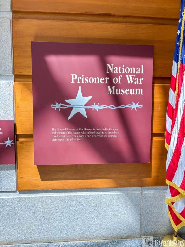 a maroon with text and a design featuring a large silver star intersected by a length of barbed wire. To the right of the panel is the edge of a U.S. flag, displaying portions of its red, white, and blue pattern with a gold fringe. The wall is bordered at the bottom by gray stone tiles.