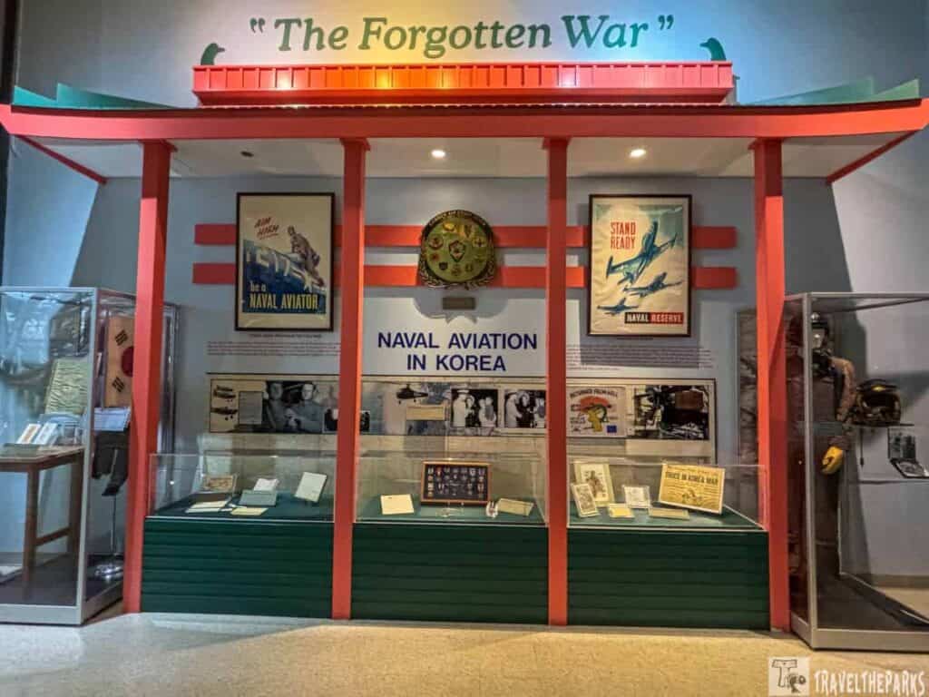 a museum exhibit titled "Naval Aviation in Korea" dedicated to the Korean War. The center of the exhibit features a sign with this title in bold blue letters. Above, in a light blue background, the words "The Forgotten War" are displayed in green font. The exhibit consists of a wall with multiple framed photographs and posters. On the left, a vintage recruiting poster displays an aviator. In the center, a collection of naval insignia and decorations are showcased in a glass case. The right side of the exhibit includes another recruiting poster featuring aircraft and figures. Three glass display cases hold various memorabilia, papers, and vintage items associated with the Korean War. The exhibit is partitioned with vibrant red columns, and the ceiling structure is adorned with red and white horizontal stripes. A mannequin wearing a flight suit and helmet stands inside a glass case on the far right.