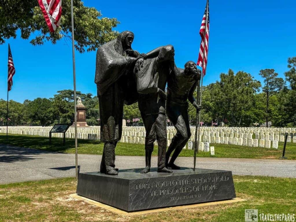 The Georgia Memorial depicts a bronze statue at Andersonville National Historic Site. The sculpture consists of three human figures in a poignant pose, embodying themes of suffering and resilience. The figure on the left drapes a cloth over the middle figure, who appears to lean on them for support. The figure on the right, standing slightly bent, holds a stick. The statue is elevated on a rectangular stone pedestal. Surrounding the statue are two American flags on tall poles, one on each side. In the background, a cemetery with aligned, rectangular headstones stretches across the landscape, set against a backdrop of lush green trees under a clear blue sky. A historical marker board and a monument are visible further in the background.
