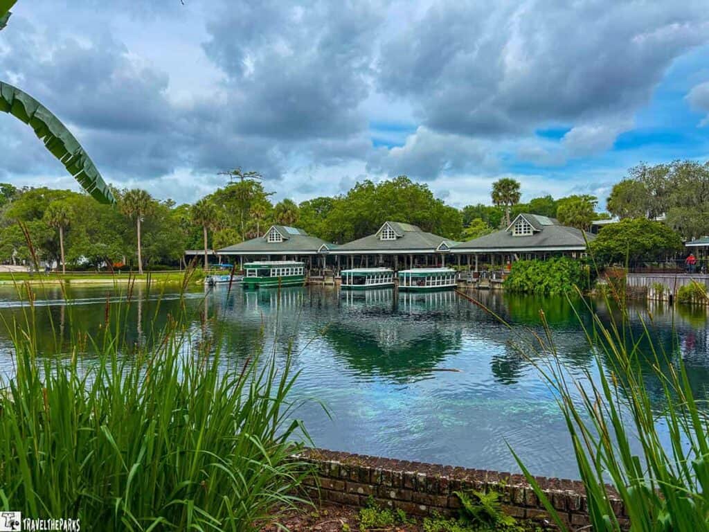 A row of buildings with green roofs by the water at Silver Springs State Park, surrounded by trees and boats, under a cloudy sky.