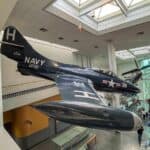 a vintage Navy aircraft suspended from the ceiling within a museum. The aircraft is dark blue with markings in white, including a large "H" on the tail, the word "NAVY," and "VF151." The aircraft has a sleek, aerodynamic design with visible cockpit windows and a sharp nose. The museum interior features a high ceiling, large skylights, and glass walls that provide natural lighting. Other aircraft and exhibit pieces are visible in the background, including a large propeller display.