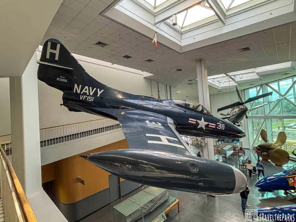 a vintage Navy aircraft suspended from the ceiling within a museum. The aircraft is dark blue with markings in white, including a large "H" on the tail, the word "NAVY," and "VF151." The aircraft has a sleek, aerodynamic design with visible cockpit windows and a sharp nose. The museum interior features a high ceiling, large skylights, and glass walls that provide natural lighting. Other aircraft and exhibit pieces are visible in the background, including a large propeller display.