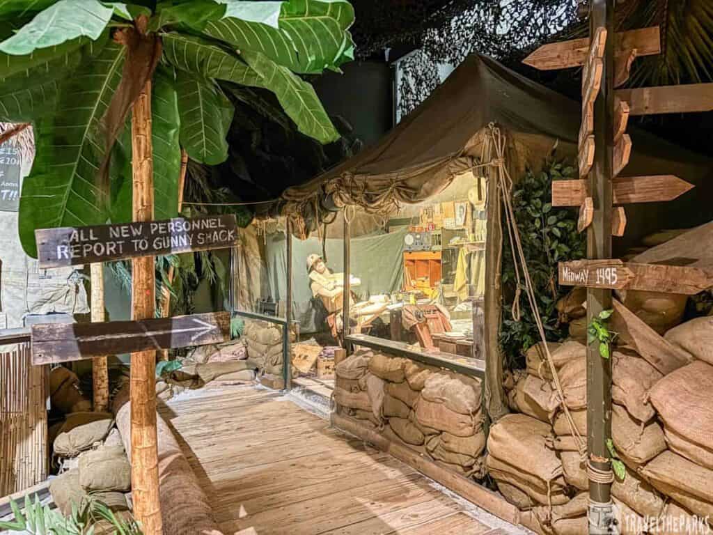  a museum exhibit depicting a military encampment at Guadalcanal. The setting includes a tent made of beige canvas material with sandbags forming a perimeter around it. Inside the tent, a mannequin dressed in military attire is seated at a desk, surrounded by vintage equipment and maps, evoking a wartime command post. To the left, a sign post made of dark wood points in various directions, and one sign reads, "ALL NEW PERSONNEL REPORT TO GUNNY SHACK" in bold letters. A tall palm tree with lush green leaves stands nearby, enhancing the realistic jungle atmosphere. The flooring consists of wooden planks, and additional sandbags line the area, reinforcing the theme of a temporary military base.