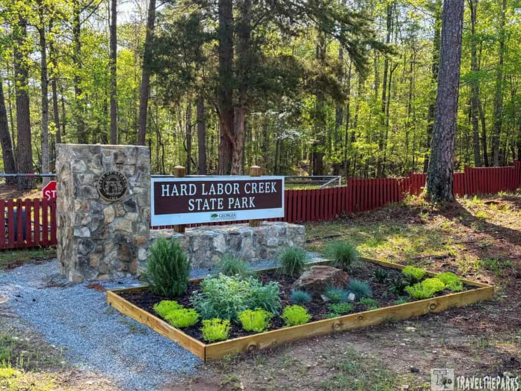 Entrance sign for Hard Labor Creek State Park with a stone column and wooden framed garden bed, set against a forested backdrop.