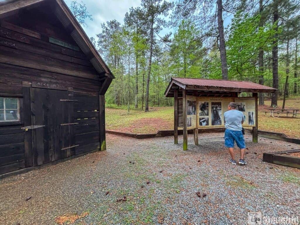 a scene at Hard Labor Creek State Park featuring a rustic, wooden structure on the left, identified as a blacksmith shop. The building is dark with visible wooden planks and a small window near the door, which has