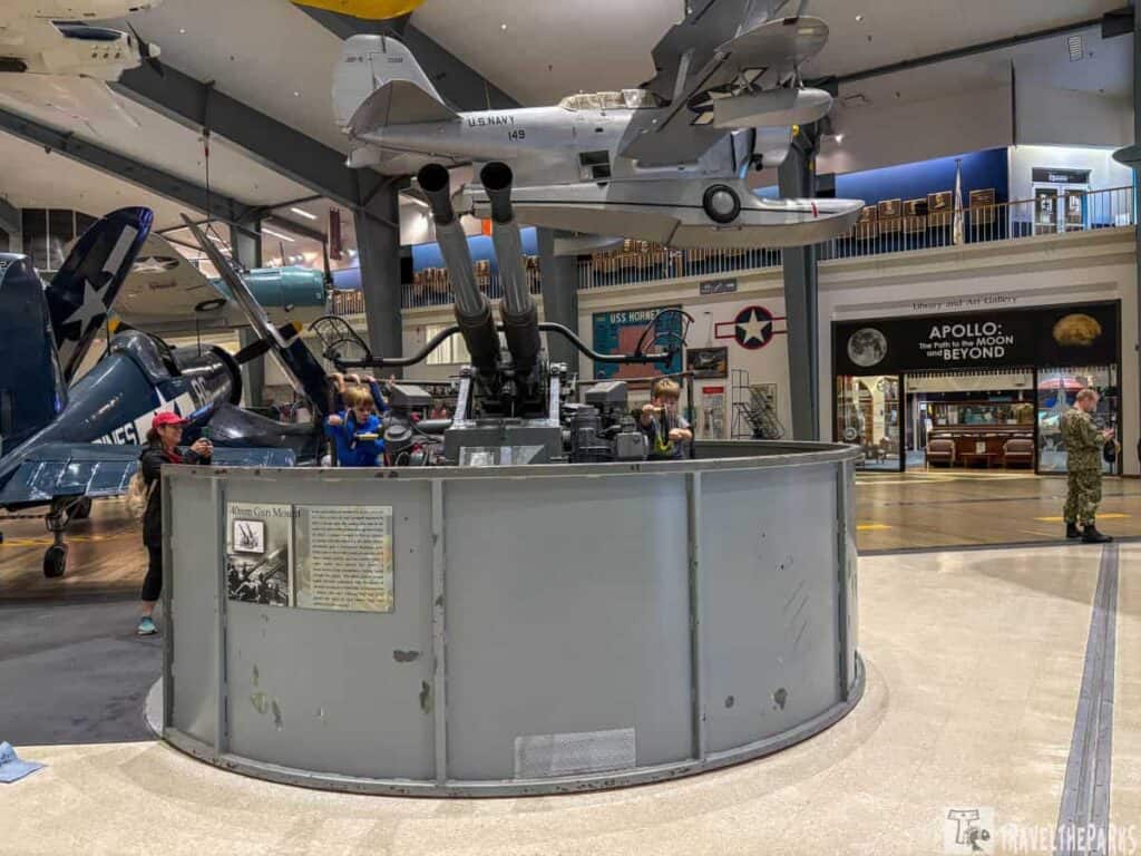 Museum exhibit featuring a 40mm gun mount and military aircraft, with visitors interacting with the displays.