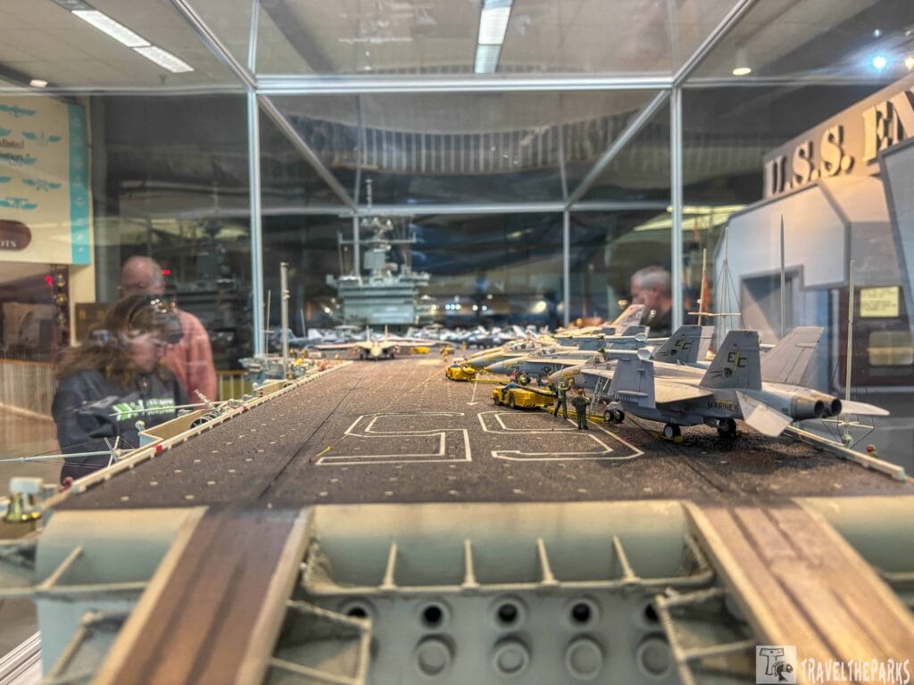 Naval Aviation Museum Pensacola a detailed diorama of an aircraft carrier within a glass case, located in a museum setting. The diorama features a scale model of an aircraft carrier deck with several fighter jets lined up on the right side, each marked with distinct insignia and numbers. In the background, a model control tower and various aircraft equipment are visible. The surface of the deck is textured, with markings indicating landing areas. The surrounding environment is visible through the glass, showing museum visitors observing the display. Overhead lighting casts reflections on the glass, adding depth to the scene.