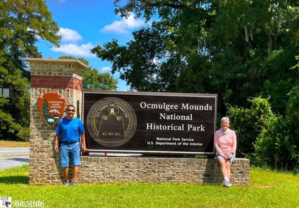 Two people near the Ocmulgee Mounds National Historical Park sign surrounded by lush trees.