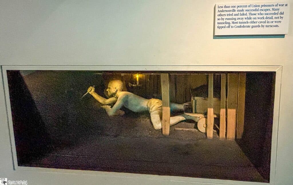Andersonville POW museum exhibit of a narrow underground tunnel. Inside the tunnel, a life-sized figure of a prisoner is shown crouched and crawling with one arm extended forward, holding a tool. The figure appears emaciated and determined, illuminated by a subtle light that casts shadows around him. The tunnel is constructed with wooden supports and has a gritty, earthen texture to emphasize its confined and rugged environment.