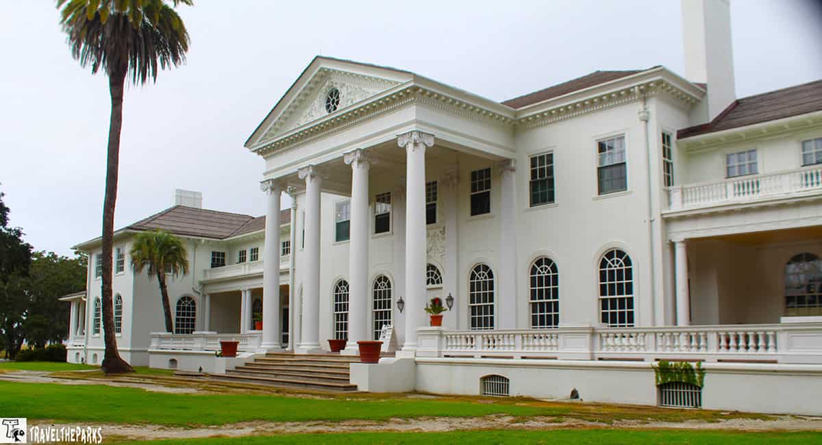 a large, elegant Plum Orchard mansion with classic architectural features. The building is predominantly white with a symmetrical facade. It is characterized by a prominent, central portico supported by several tall, white columns that lead up to a pediment adorned with decorative detailing. The mansion has multiple windows with white sashes, some arched and some rectangular, lined along the front. A wide set of steps leads up to the main entrance. The roof is slightly pitched and features chimneys on both ends. There are large palm trees and well-manicured grass visible in the foreground, contributing to the stately appearance of the property.