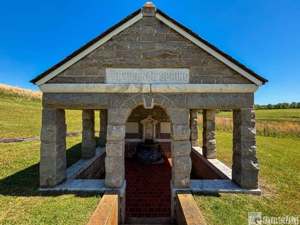 open-air stone structure known as Providence Spring at Andersonville National Historic Site. The rectangular building features a gabled roof with shingles and is supported by four stone pillars. The front of the structure has an arched opening, revealing an interior lined with red tiles on the floor and a stone fountain centered inside. Sitting atop the gable is a horizontal stone sign with the words "Providence Spring" engraved into it. The surrounding landscape consists of rolling grassy hills under a clear blue sky, suggesting a peaceful and open environment.