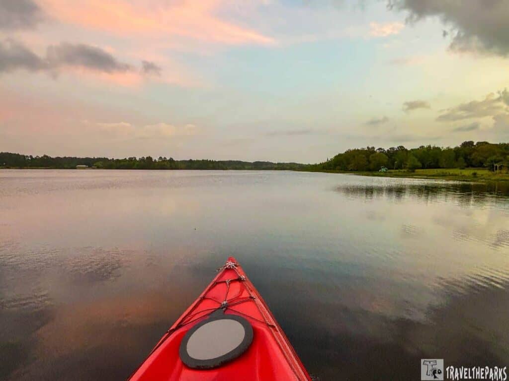 lake at Reed Bingham State Park during what appears to be dawn or dusk. In the foreground is the nose of a red kayak, partially visible. The kayak is positioned in the calm waters, pointing towards the horizon. The water reflects the soft pastel hues of the sky, which is painted in gentle shades of pink, orange, and blue. To the right, the shoreline is lined with a dense treeline, showing lush greenery. The distant shore stretches across the image, with trees and possibly other brush visible. The scene evokes a sense of tranquility and natural beauty that is typical of lakeside landscapes.