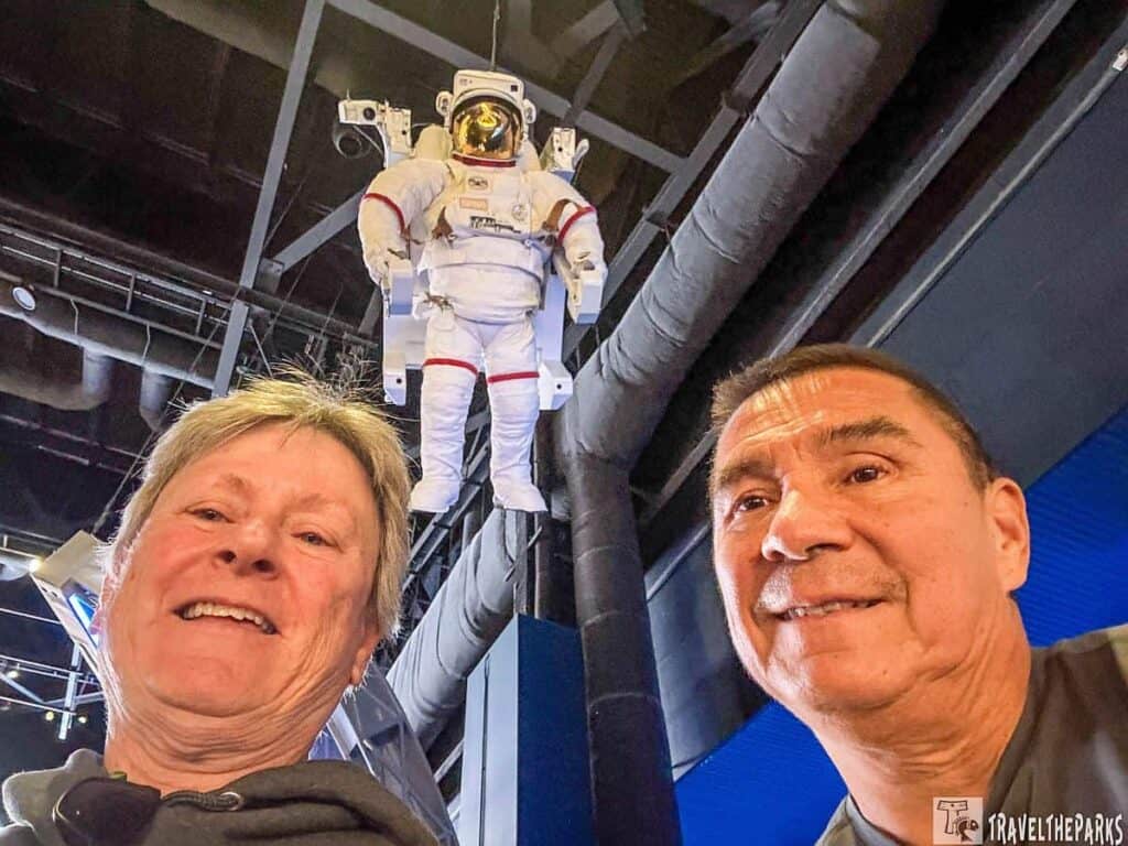 Two people taking a selfie with a suspended astronaut model above them.