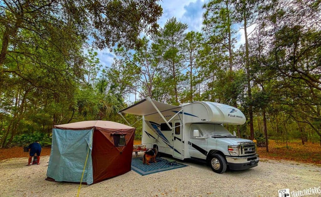 RV and canopy setup at a wooded campsite with a dog under the RV's awning.