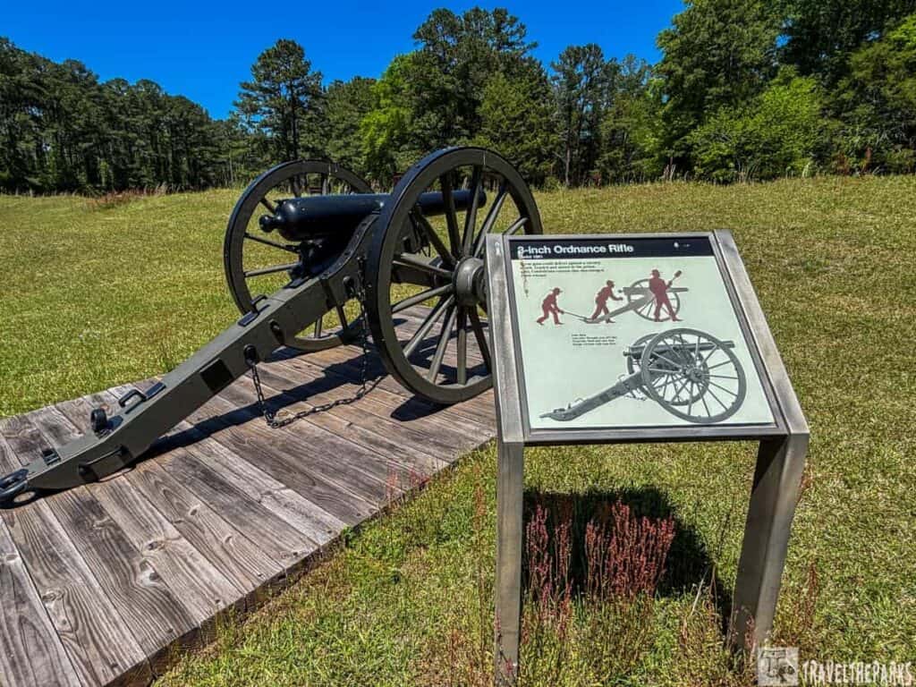 historical cannon, identified as a 3-inch Ordnance Rifle, positioned on a wooden platform in a grassy area. The cannon is black with large spoked wheels and a long barrel. Adjacent to the cannon is an informative sign mounted on a metal stand, featuring an illustration of the cannon and text explaining its use. In the background, a line of dense trees borders the open field under a clear blue sky.