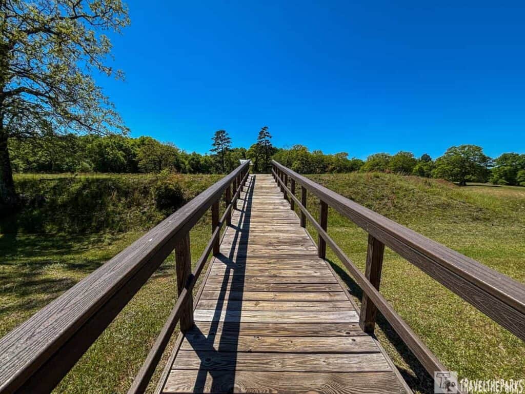 A wooden boardwalk extending into the distance, framed by sturdy railings on both sides. The planks of the boardwalk are a natural wood color, and they stretch forward toward a horizon filled with lush greenery and tall trees. The sky is clear and vibrant blue, contrasting with the greens of the surrounding landscape. Shadows from the railings create a pattern on the wooden walkway. On the left, a large tree extends its branches, adding depth to the scene. The ground on either side of the boardwalk is grassy, appearing well-tended and slightly sloped. A wooden boardwalk extending into the distance, framed by sturdy railings on both sides. The planks of the boardwalk are a natural wood color, and they stretch forward toward a horizon filled with lush greenery and tall trees. The sky is clear and vibrant blue, contrasting with the greens of the surrounding landscape. Shadows from the railings create a pattern on the wooden walkway. On the left, a large tree extends its branches, adding depth to the scene. The ground on either side of the boardwalk is grassy, appearing well-tended and slightly sloped.