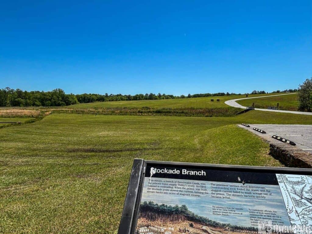 Andersonville National Historic Site. Rolling grassy fields dominate the foreground, gradually sloping upwards toward the horizon. A paved path winds through the scene, leading to a vanishing point in the distance. Surrounding the fields are scattered trees and shrubs, framing the open space with verdant foliage. In the lower right corner, a large informational sign titled "Stockade Branch" is visible, partially detailing the historical context of the area. The sign includes a text section and a historical illustration.