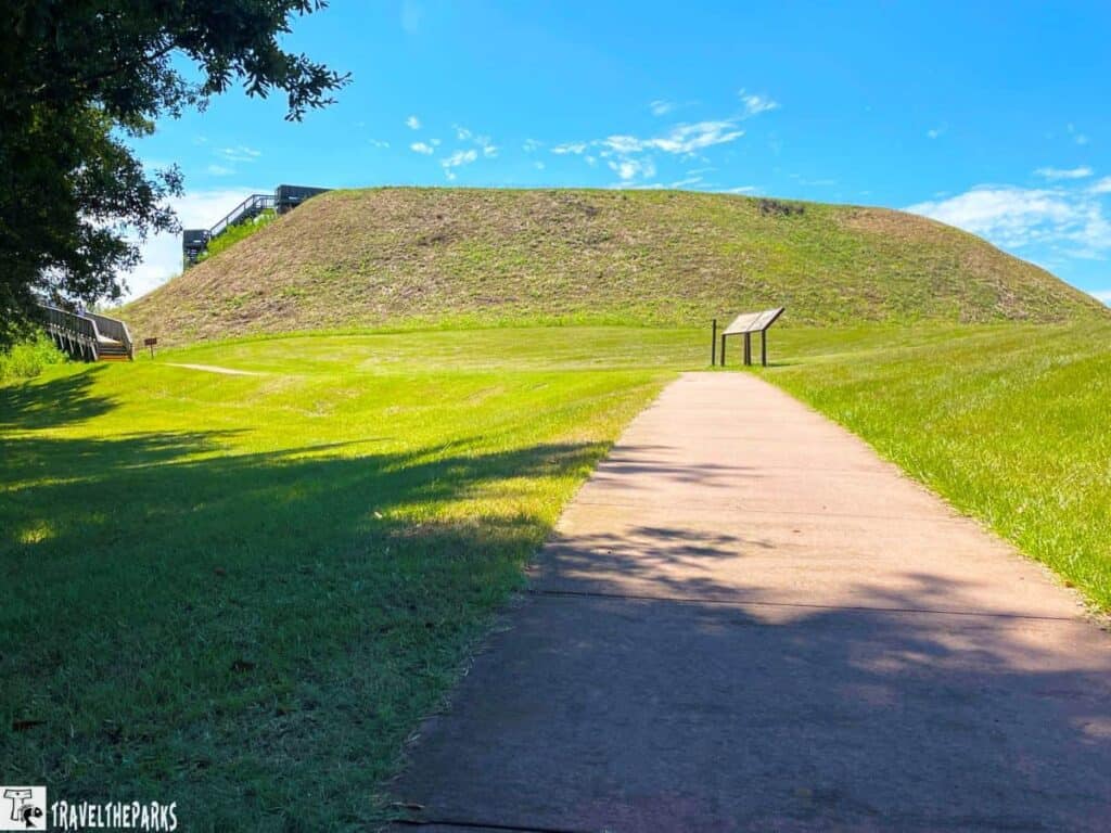 A paved path leading to a large, grassy mound with signs and an adjacent staircase under a blue sky.