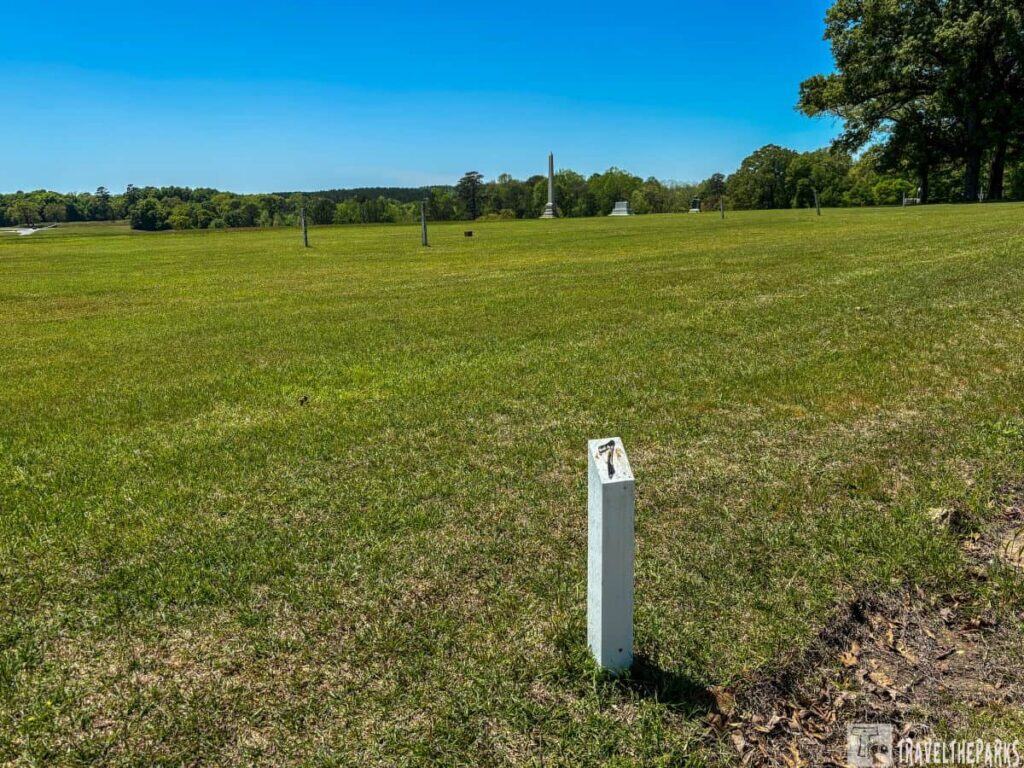 In the foreground, a white rectangular post is embedded in the ground. The grass is lush and green, extending into the background where trees border the field, providing a natural backdrop. Scattered across the field are some standing structures and monuments, visible as tall, narrow shapes. The clarity of the sky and the vibrant color of the grass suggest clear weather, and the shadows hint at midday sunlight.