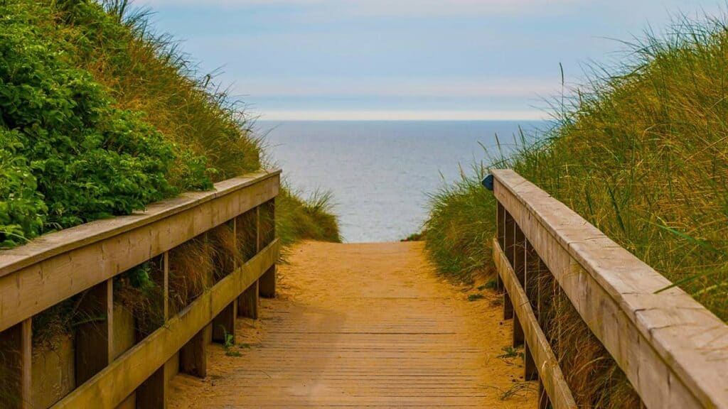 A sandy boardwalk through grassy dunes leads to the ocean under a blue sky along the Emerald Coast of Florida.
