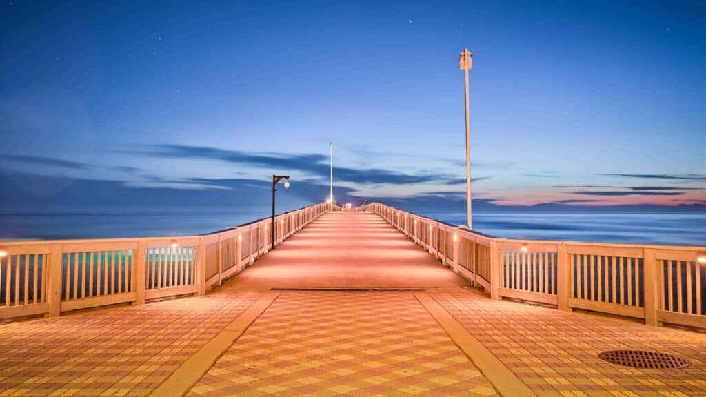 A boardwalk over the ocean at twilight, with wooden railings and lights, under a blue sky with scattered clouds.