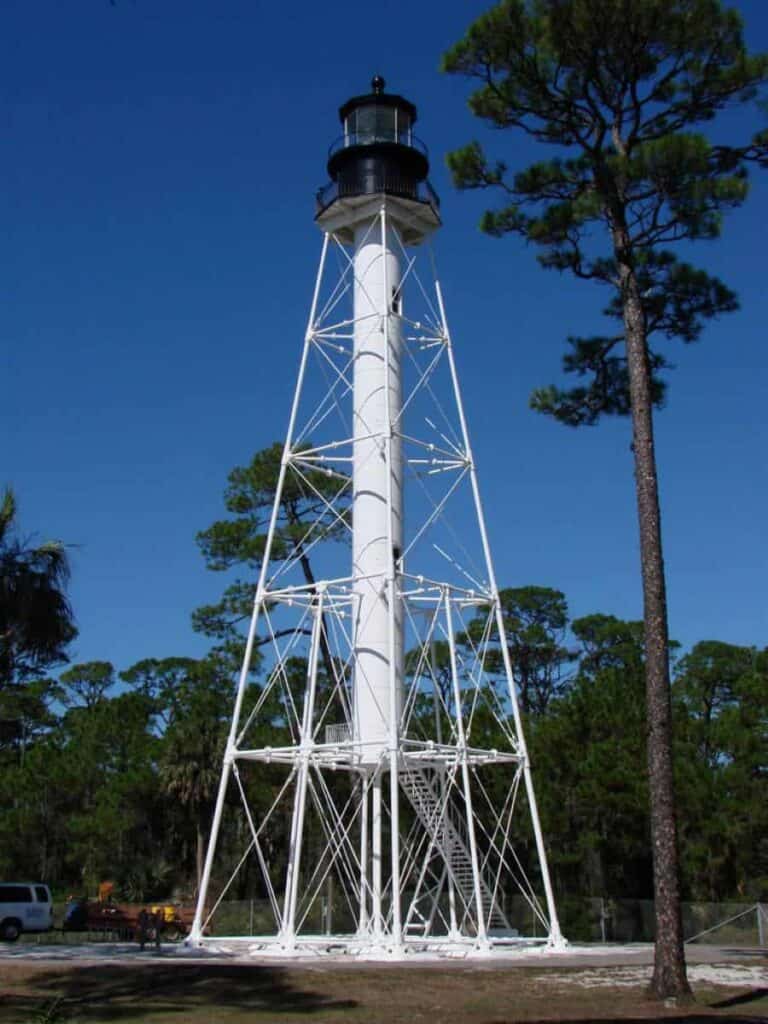 A tall, skeletal white lighthouse set against a bright blue sky. The SAn Blas lighthouse on the forgotten coast features a black lantern room at the top, with a circular viewing platform just beneath it. The structure consists of a central cylindrical column surrounded by a network of supporting white metal beams that form a lattice tower. At the base, a small set of stairs leads up to the entrance. The lighthouse is situated in a natural setting, with surrounding greenery including pine trees and various shrubs. The overall setting suggests a coastal environment, enhanced by the clear blue sky and lush vegetation. FGP