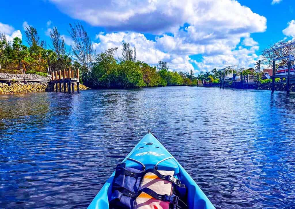 View from a blue kayak on a calm river with trees and docks on either side under a blue sky.