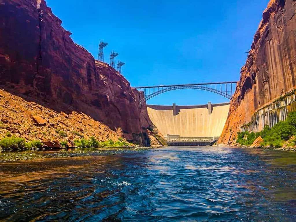 View of a dam and arch bridge at Glen Canyon, framed by red cliffs and blue water.