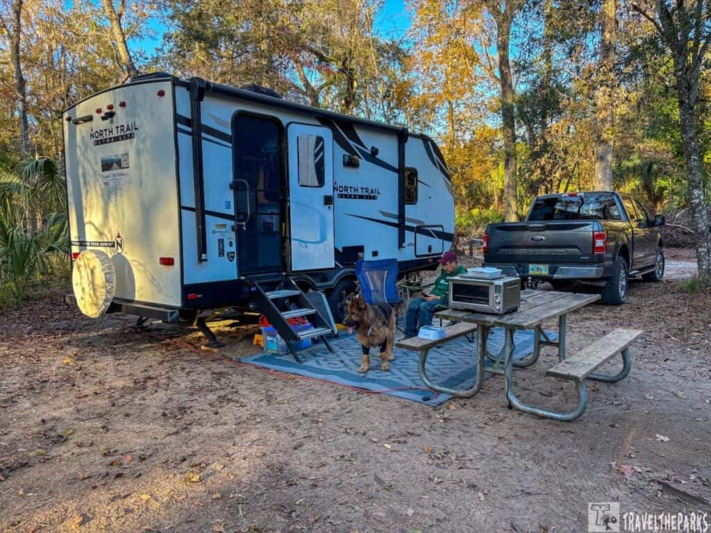 Campsite with a travel trailer, pickup truck, and picnic setup in a wooded area.