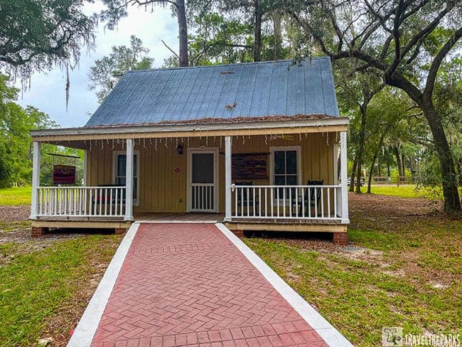 A small beige cabin with a metal A-frame roof and a brick pathway leading to its porch.