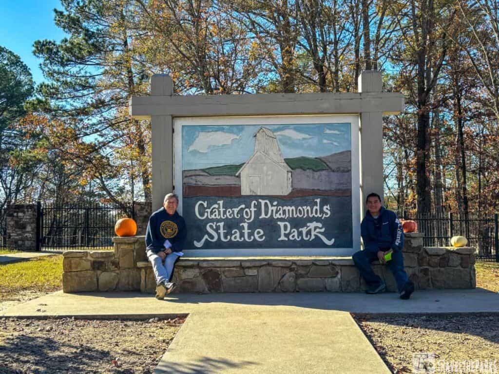 Sign for Crater of Diamonds State Park with a painted mural and two people seated in front.