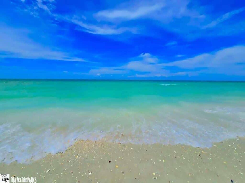 A tropical beach with turquoise water, small waves, and a sandy shore under a bright blue sky with white clouds.