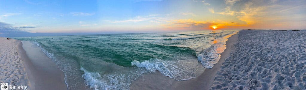  a panoramic view of a beach on the Emerald Coast during sunset. The shoreline curves gently from the left foreground towards the horizon on the right. The sand is a light, almost white color, textured with footprints. The ocean displays varying shades of green and blue, with small waves gently lapping against the shore. In the distance, the sun sets, casting vivid shades of orange and yellow across the sky and reflecting on the water's surface. The sky transitions from the warm hues of the sunset to cooler blues 