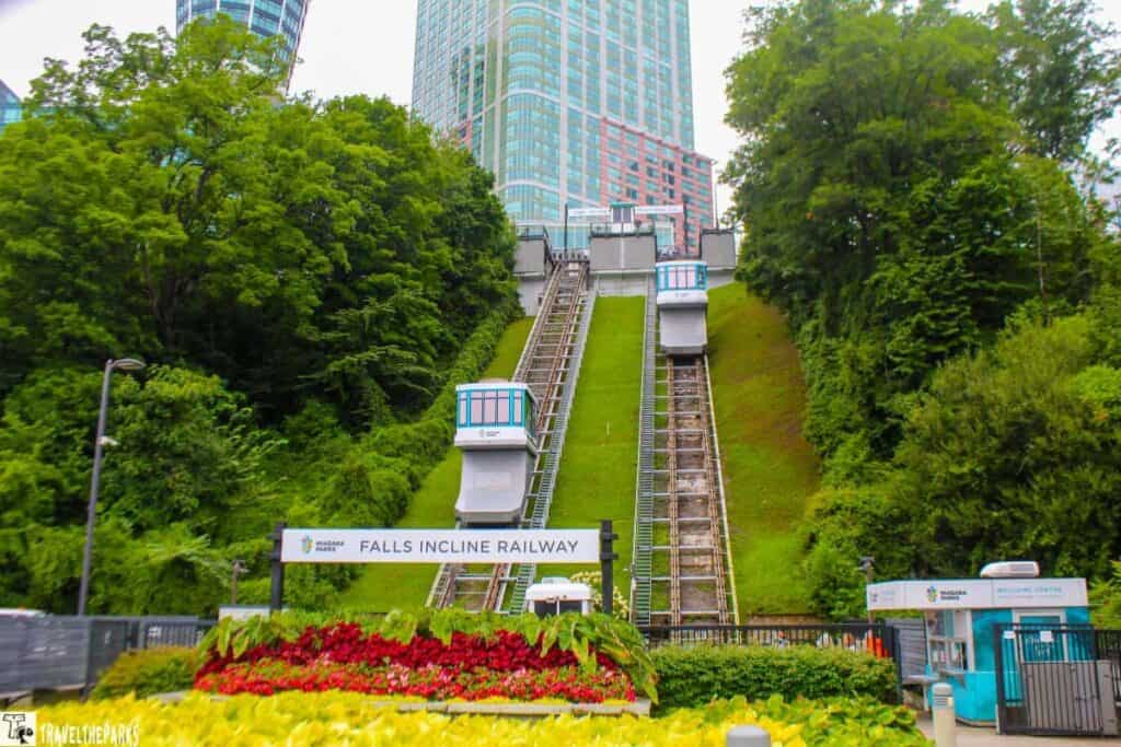 Incline railway with two tracks and small trams on a grassy slope, surrounded by trees and tall buildings in the background.