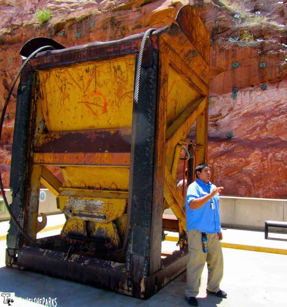 A rusted yellow industrial concrete bucket with a person in a blue shirt gesturing, set against a red rock wall.