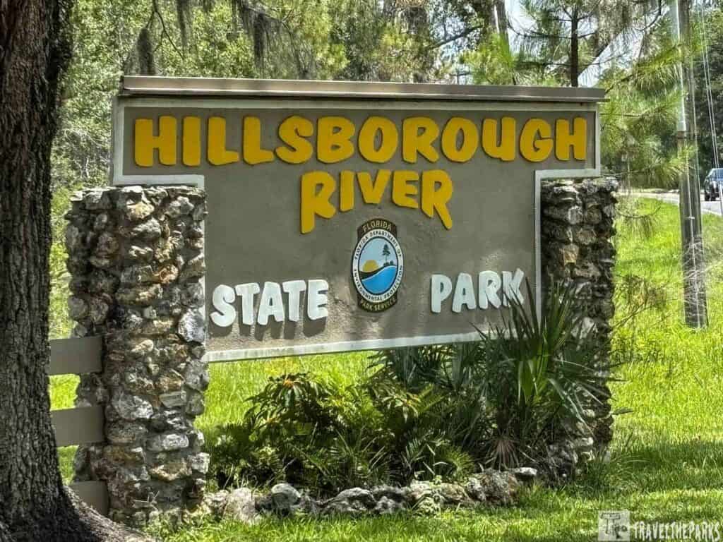 park entrance sign for Hillsborough River State Park. The sign is primarily rectangular and composed of a beige background bordered by stacked stones. In bold, uppercase letters, "Hillsborough River" is written in yellow at the top, while "State Park" is written in white at the bottom. Between the text, a circular emblem featuring the words "Florida State Parks," with a small graphic of a palm tree and water, is visible. The sign is situated in a lush outdoor environment, surrounded by greenery including grass, small plants, and tall trees providing shade.
