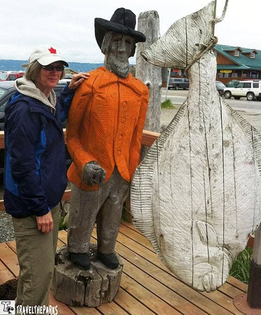 A person stands next to a wooden statue of a man and a large wooden fish sculpture on a boardwalk, with buildings and parked cars in the background.