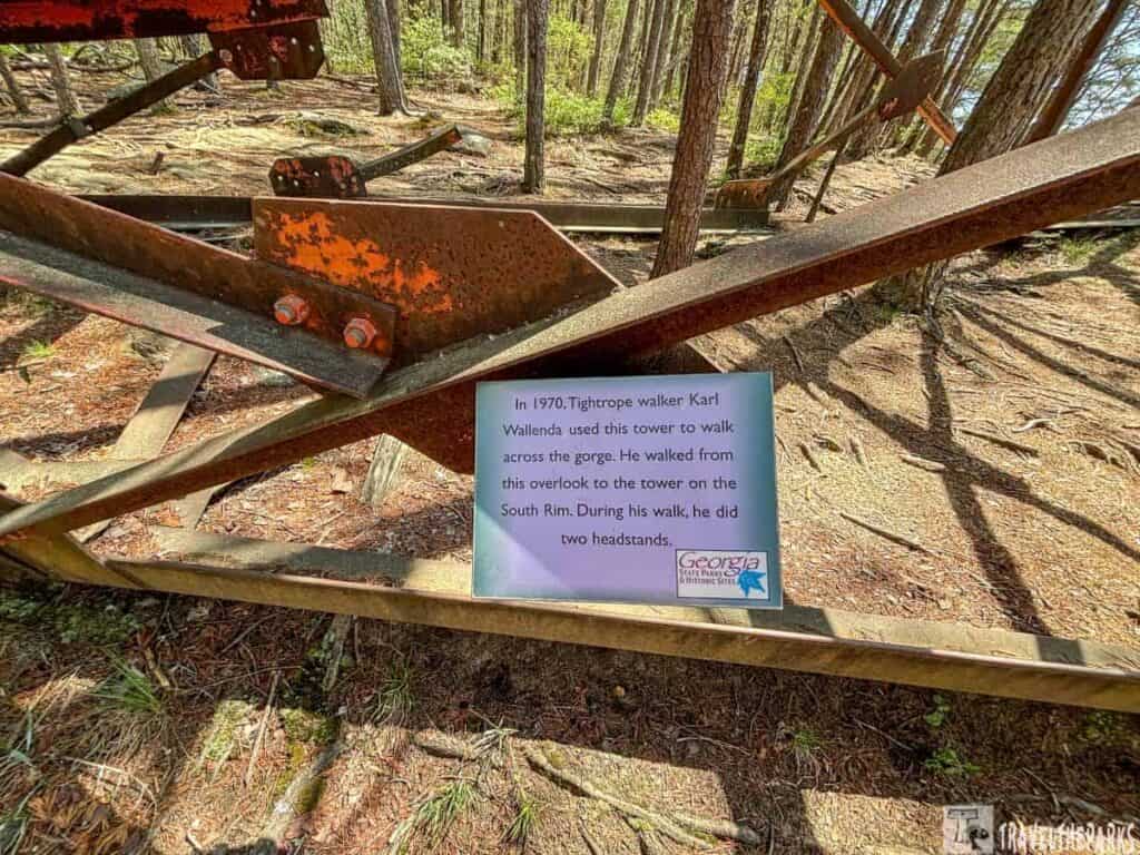 A partially rusted metal structure in a forest with a sign describing a historical tightrope walk.
Transcribed Text:
In 1970, Tightrope walker Karl Wallenda used this tower to walk across the gorge. He walked from this overlook to the tower on the South Rim. During his walk, he did two headstands. Georgia State Parks & Historic Sites.