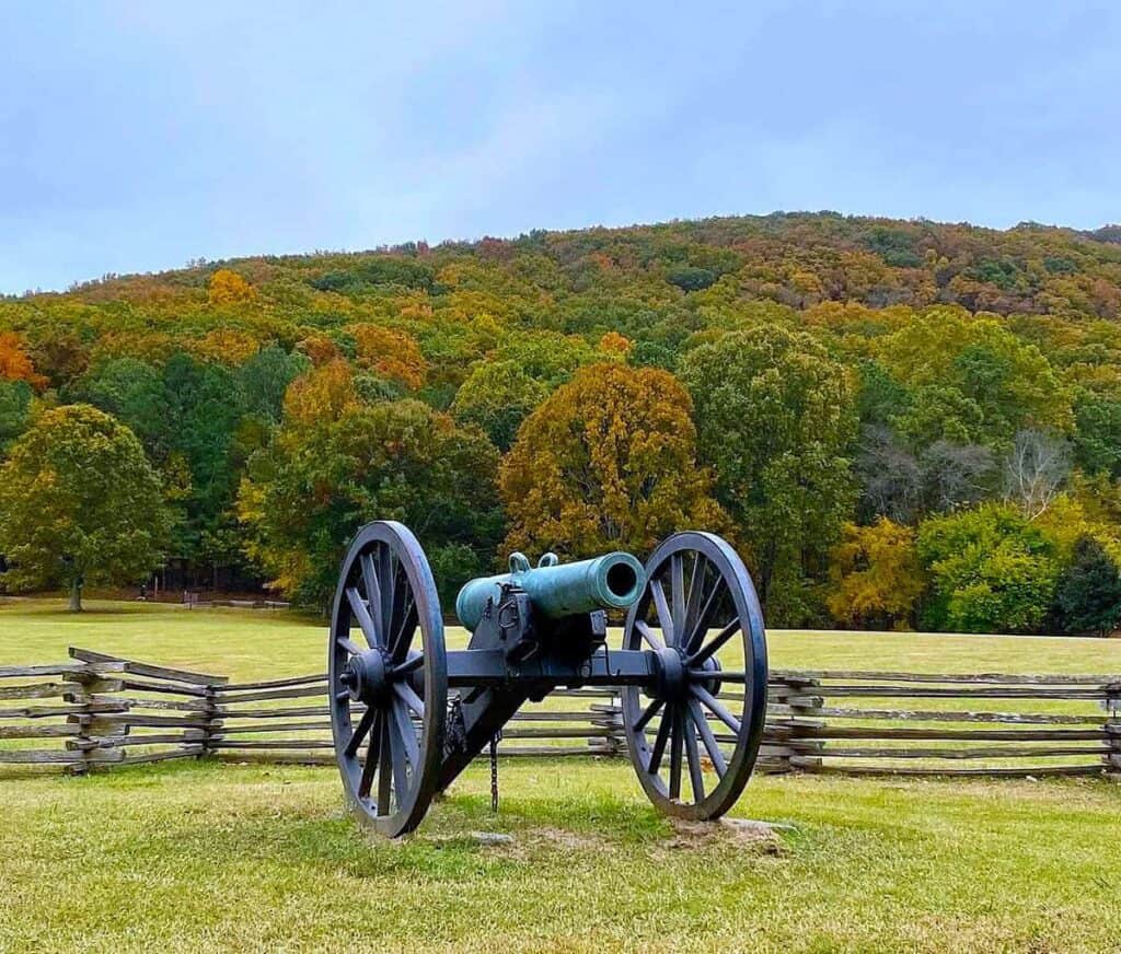 A green cannon on wooden wheels in a grassy field at Kennesaw Battlefield, with autumn trees in the background.