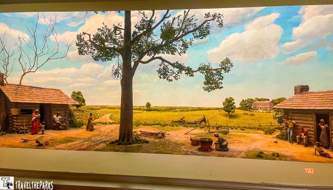 Diorama of a rural scene with cabins, people, and a large central tree under a cloudy sky.