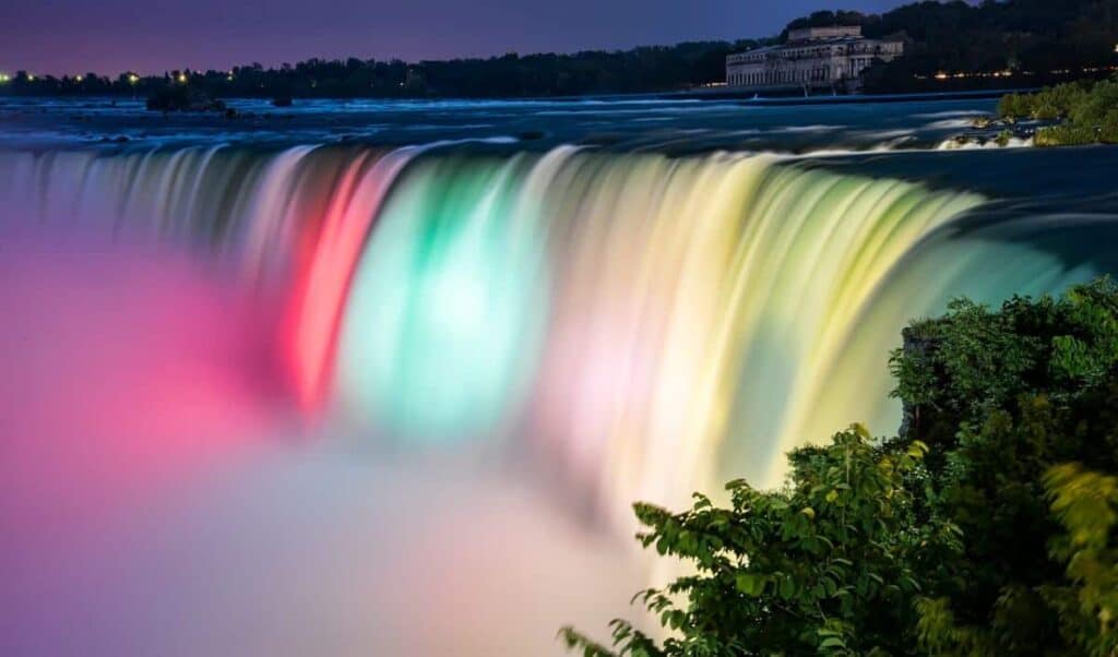 Niagara Falls at night, brightly illuminated with colorful lights. The water cascades over the edge, flowing smoothly and creating a mist at the base. Vivid colors of red, green, and yellow are projected onto the falls, creating a striking visual effect. In the background, a building with classical architecture can be seen, surrounded by dark, wooded terrain. The sky above is a deep purple-blue, contrasting with the bright, illuminated falls. In the foreground, lush green foliage adds texture and depth to the scene.