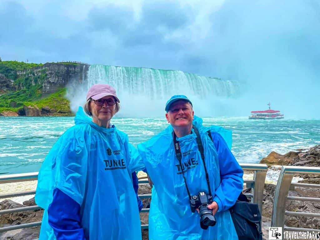 Two people in blue ponchos stand in front of Niagara Falls with a tour boat nearby.