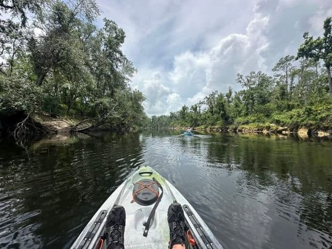 View from a kayak on a calm river with trees on both sides and another kayak in the distance.