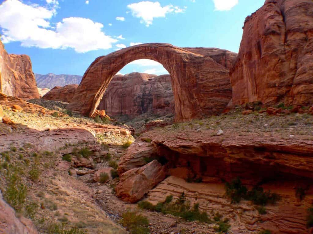 The Rainbow Bridge, a large natural rock arch in a desert canyon, with vivid red and orange hues under a bright blue sky with scattered clouds.