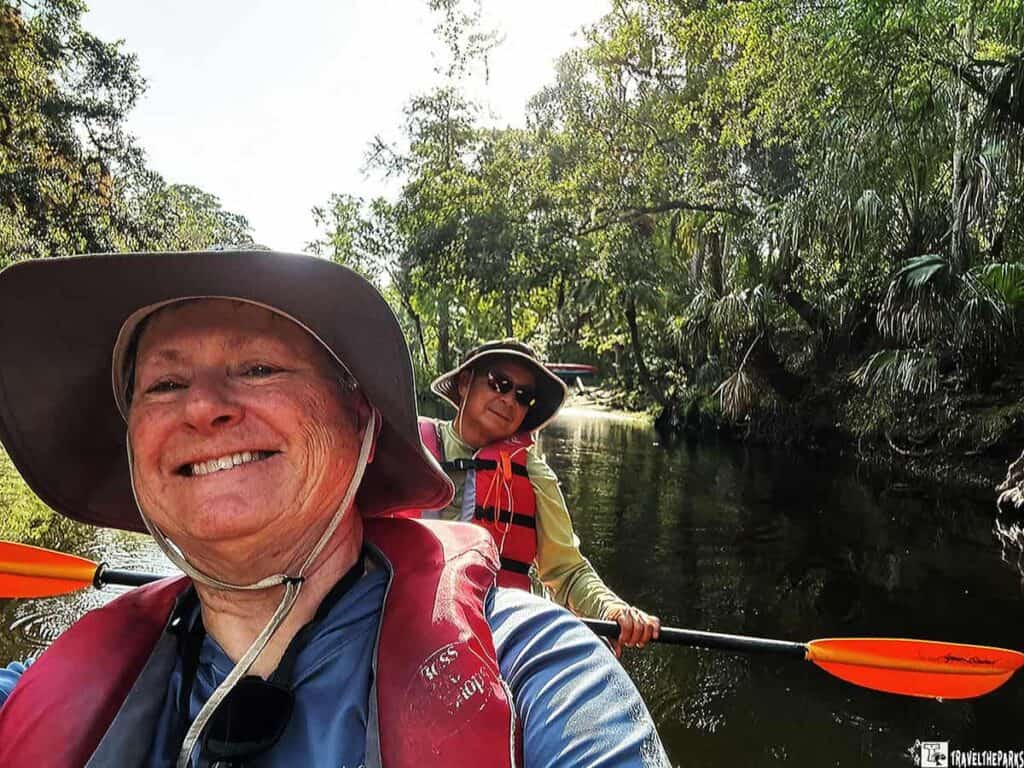 Two people kayaking on a tree-lined river, wearing hats and life jackets.