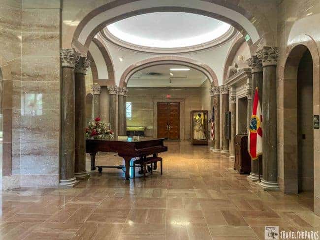 A hall with polished stone flooring and columns, featuring a grand piano and display cases.